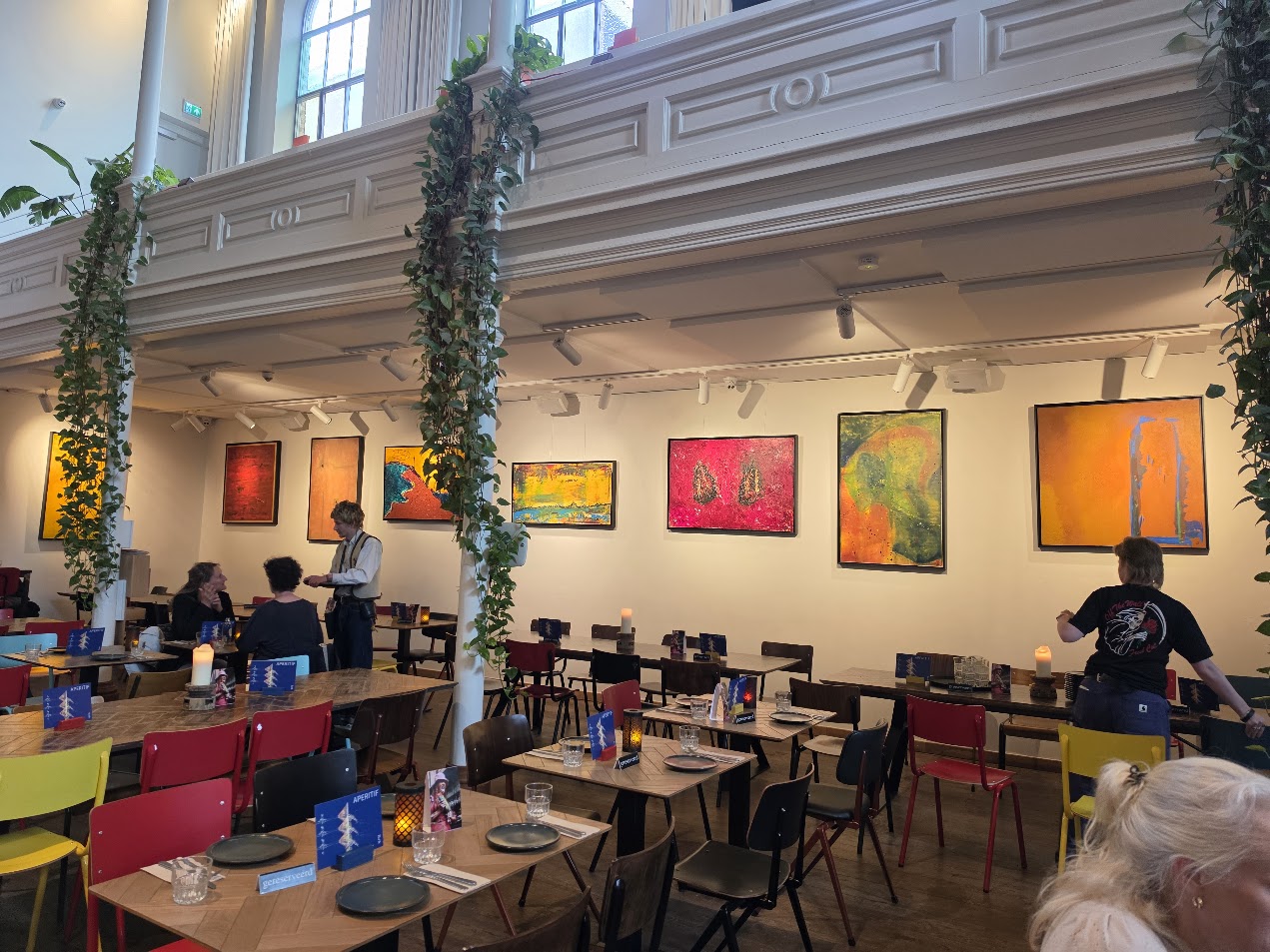 Paintings by Ward Schepman above tables at BUNK, in a tall space with columns and hanging plants.